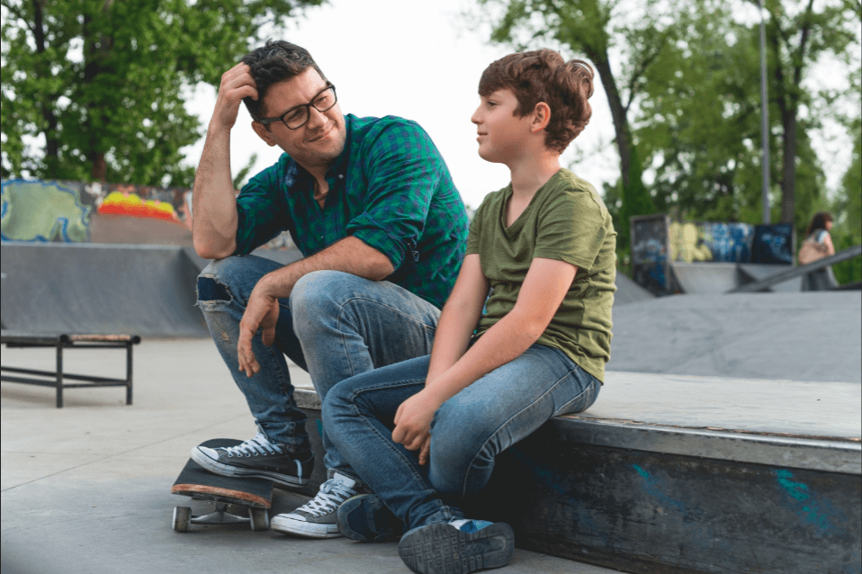 An image of a father sitting in a skate park, talking to his young son