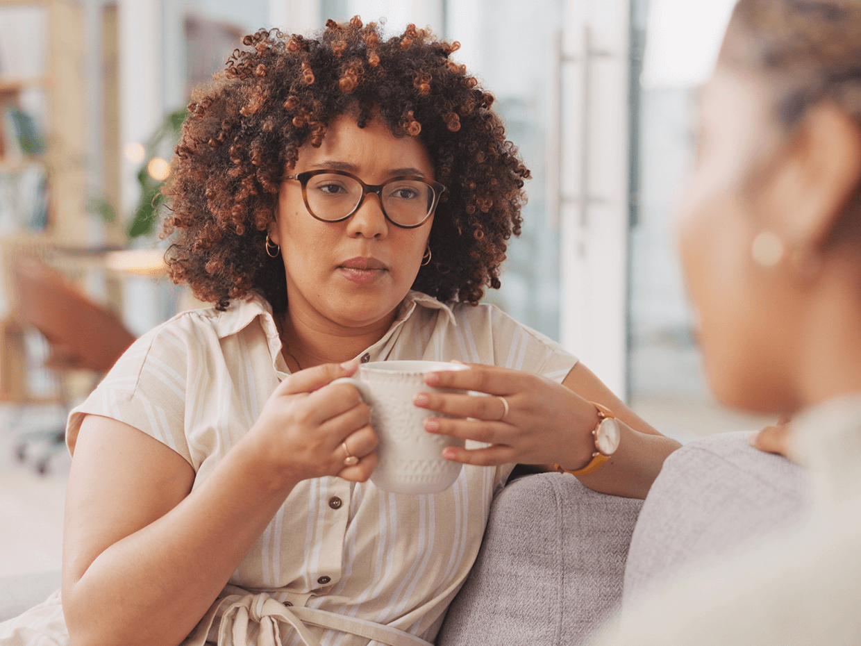 A woman with glasses and a cup of coffee sitting on a couch engaged in conversation..